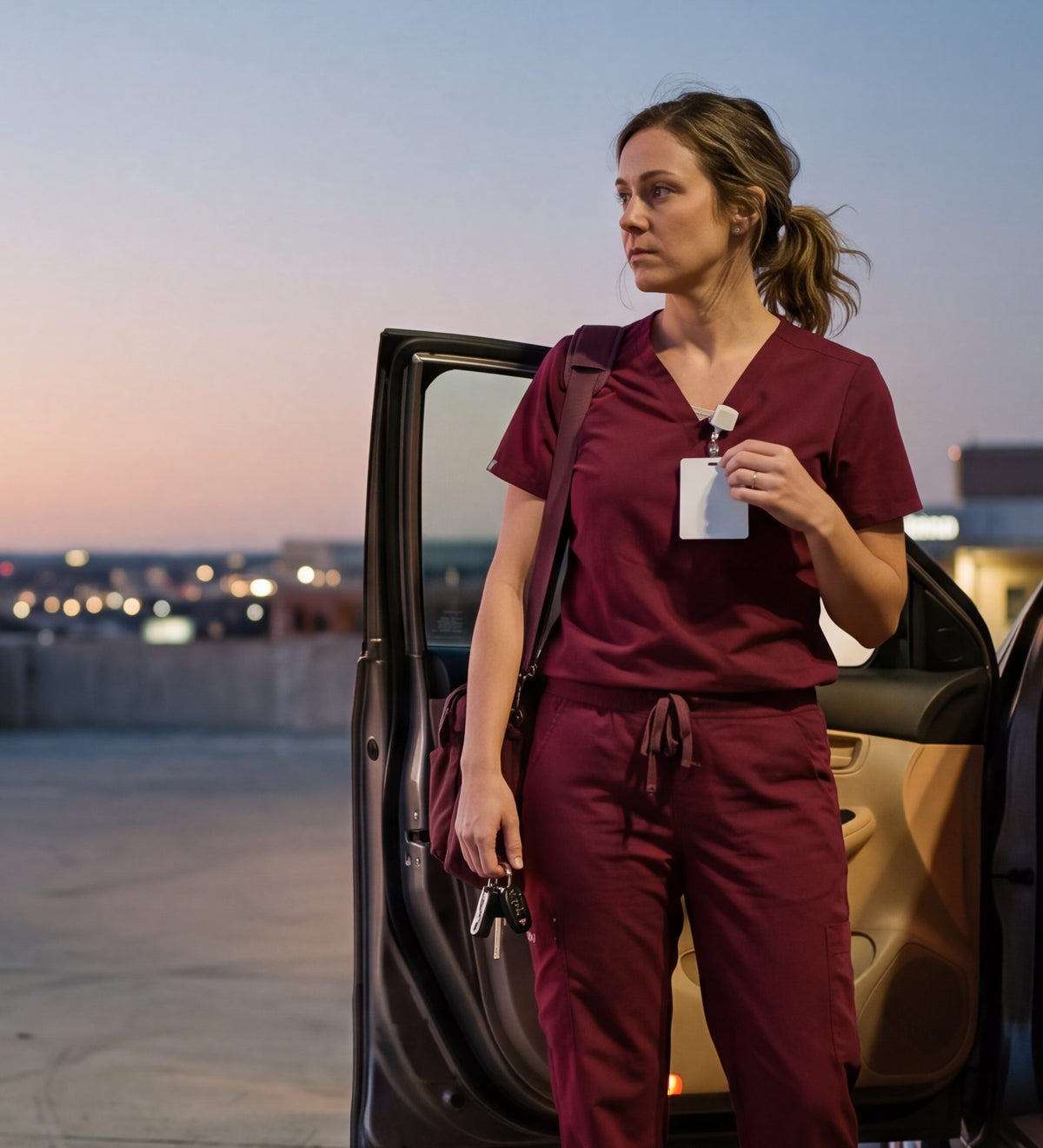Nurse in maroon scrubs standing by her open car door on a parking garage roof at dusk