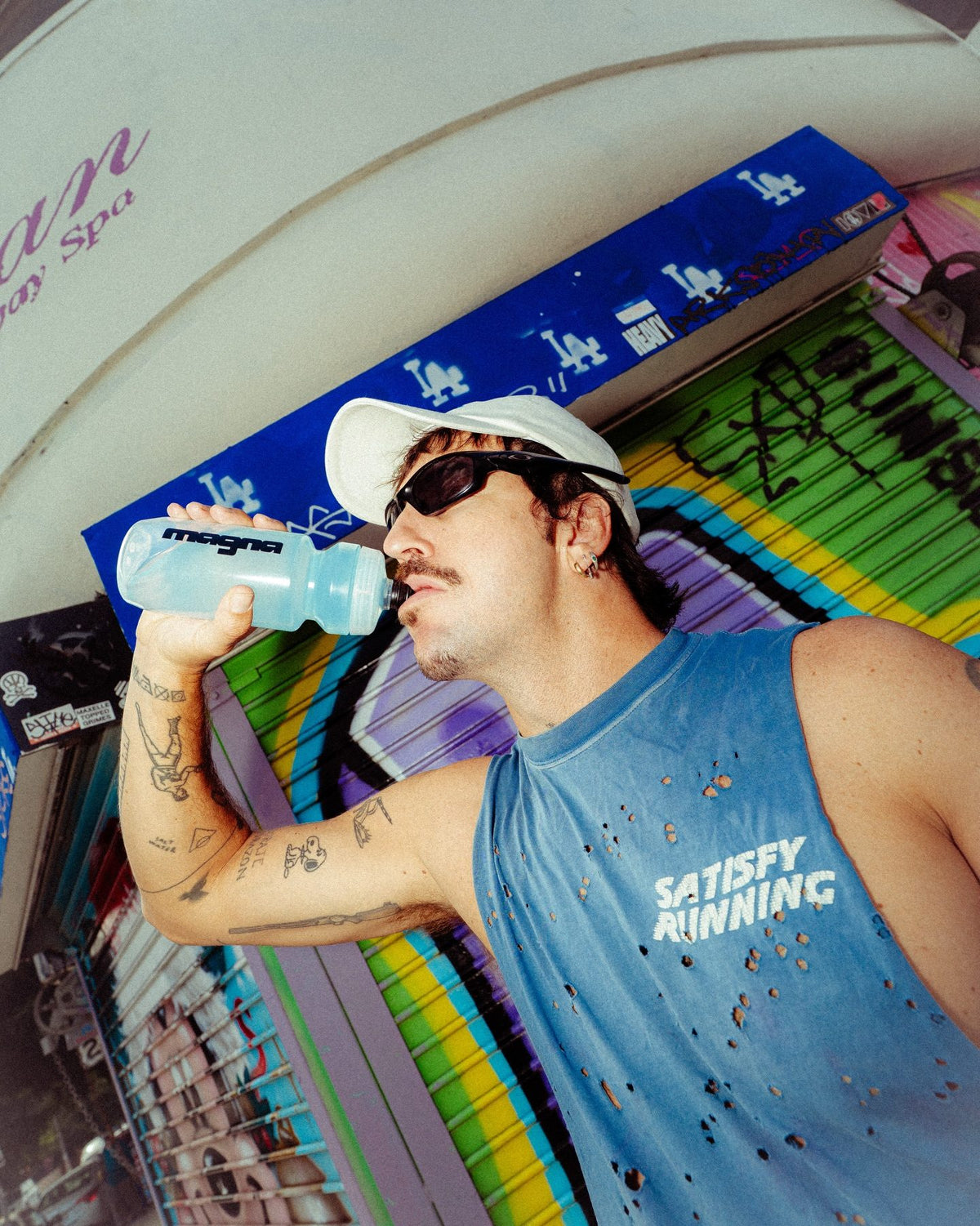 Man in a white cap and sunglasses drinking from a Magna water bottle in front of a graffiti wall
